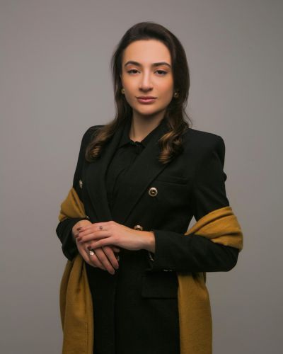 Foto de Alícia Lizz Maia Lopes Pinheiro, colaboradora no escritório de advocacia Gelson Ferrareze. Ela está sorrindo, com cabelos loiros longos soltos, vestindo uma blusa preta de gola alta e um colar dourado com um pingente de cruz. Seus braços estão cruzados, e ela usa um xale azul escuro sobre os ombros. O fundo é neutro e escuro, destacando sua imagem.
